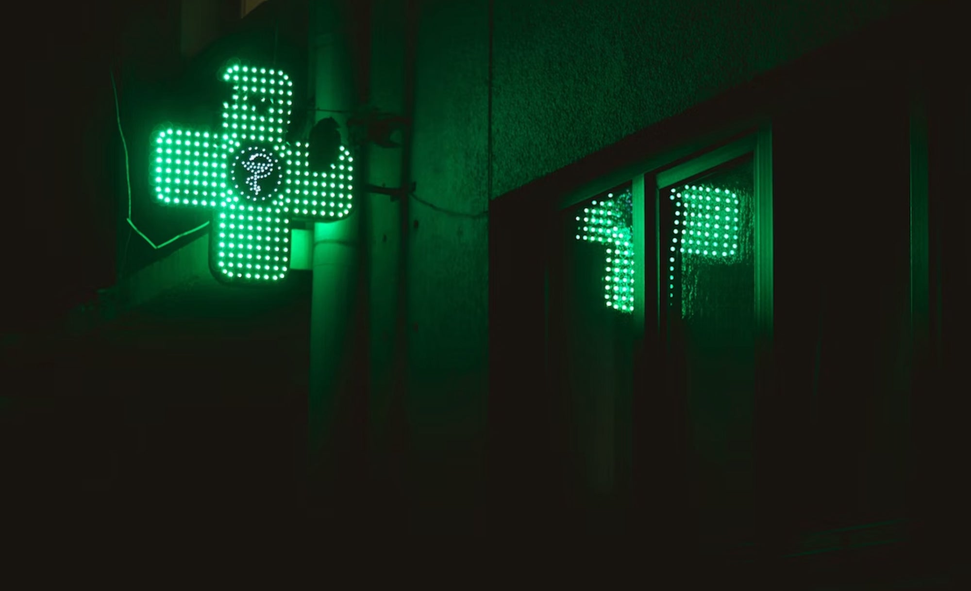 Croix de pharmacie verte lumineuse la nuit, reflétée dans une fenêtre
